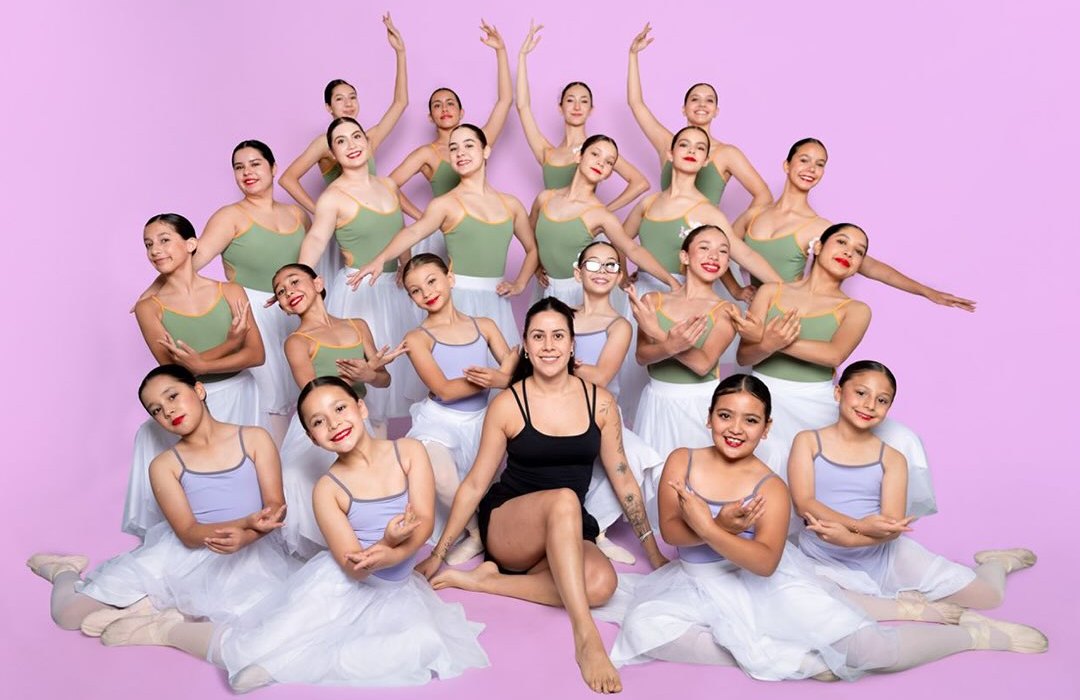 Alumnas de Ballet Brío posan juntas sobre fondo lila, con su maestra sentada al centro.